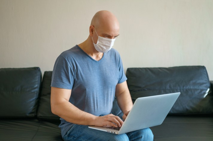 Bald man works at home on a laptop on a sofa wearing a mask during quarantine